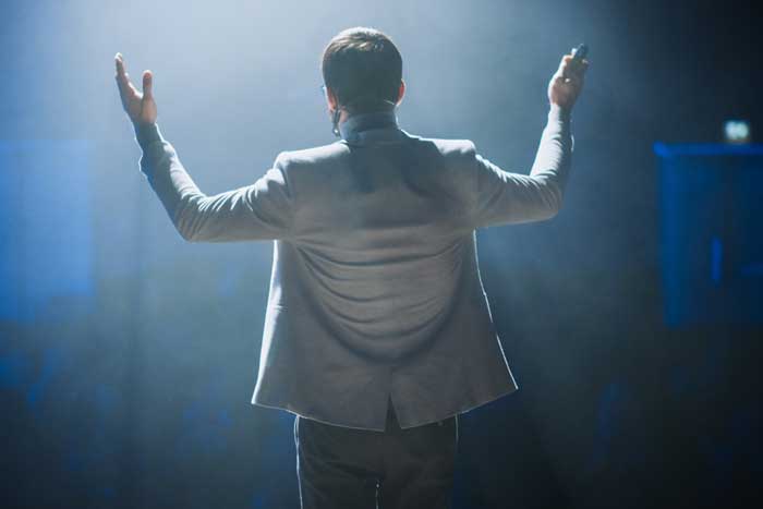 un orateur sur scène vue de dos, les bras levés, dans une ambiance bleutée